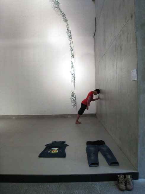 A performer leans her head and hands against the right hand wall, a set of clothes are neatly laid at the front of the space and spirally up the back wall, a series of weblike bronze sculptures can be seen