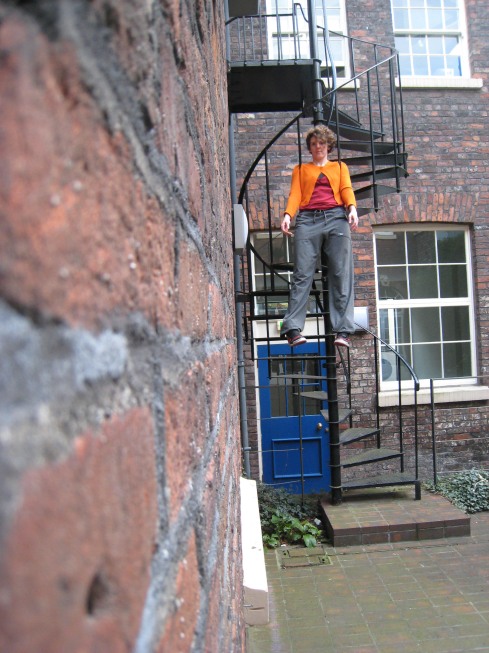 A young woman in an orange top hands from a fire escape, the railings of the stairs tucked under her arms