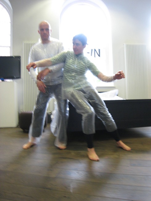 Two figures stand in front of a sunlit window in the middle of a contact improvisation. Each is wearing bubble-wrap over their everyday clothes